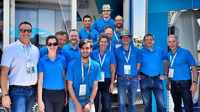 A group of ICL team members wearing blue shirts pose together outside a company booth at an outdoor event. They are smiling and appear enthusiastic, representing teamwork and collaboration.
