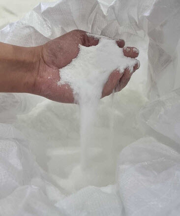A hand holding and pouring fine white granular fertilizer from a large open bag, showing the texture and purity of the material.