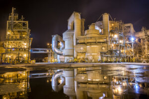 A brightly lit industrial plant at night with complex metal structures and large machinery, reflected in a puddle on the ground.