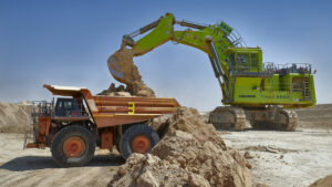 A large green excavator loads mineral-rich soil into a heavy-duty dump truck at an open-pit mine under a clear blue sky.