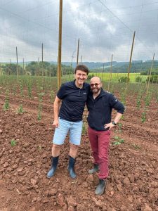 Two men smiling in a hop field with tall trellising poles.