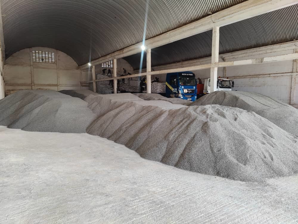 Large storage facility filled with mounds of Polysulphate fertilizer granules, showing bulk material ready for transport and agricultural distribution, with trucks and workers visible in the background.
