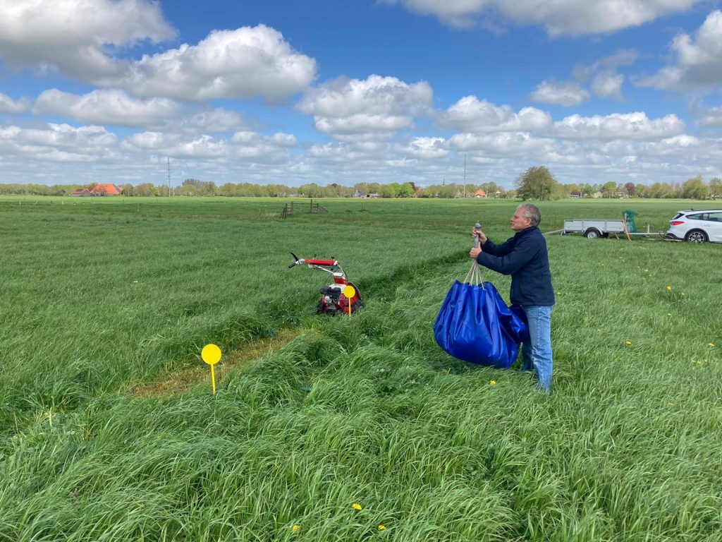 Lex Slootweg standing in a green grassland field under a partly cloudy sky, holding a large blue bag near a small red agricultural machine used for trials.