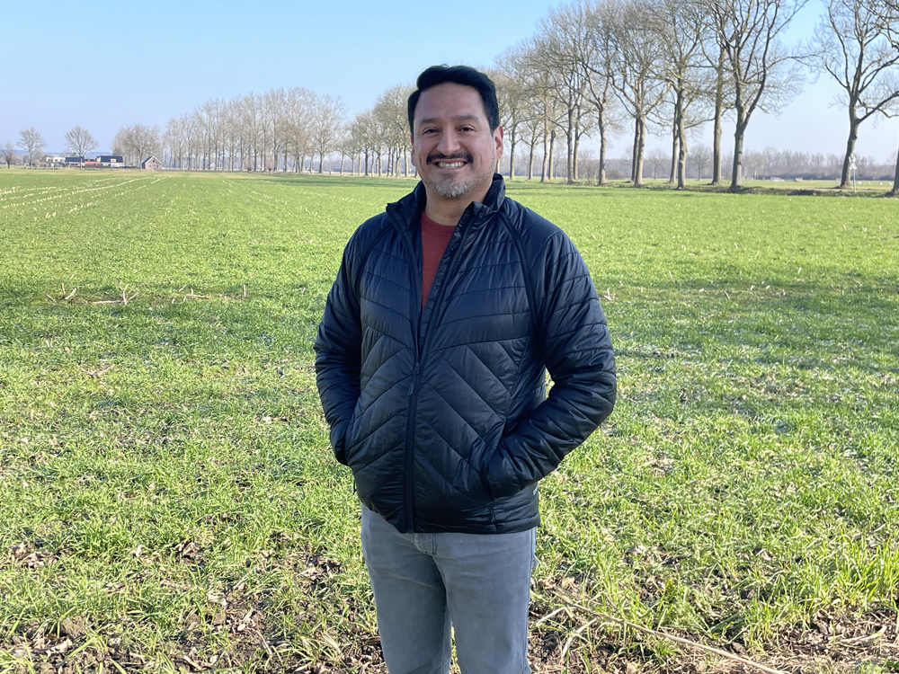 Cristian Terrones standing in a green field in the Netherlands, wearing a dark jacket and smiling on a clear, sunny day with leafless trees lining the background.