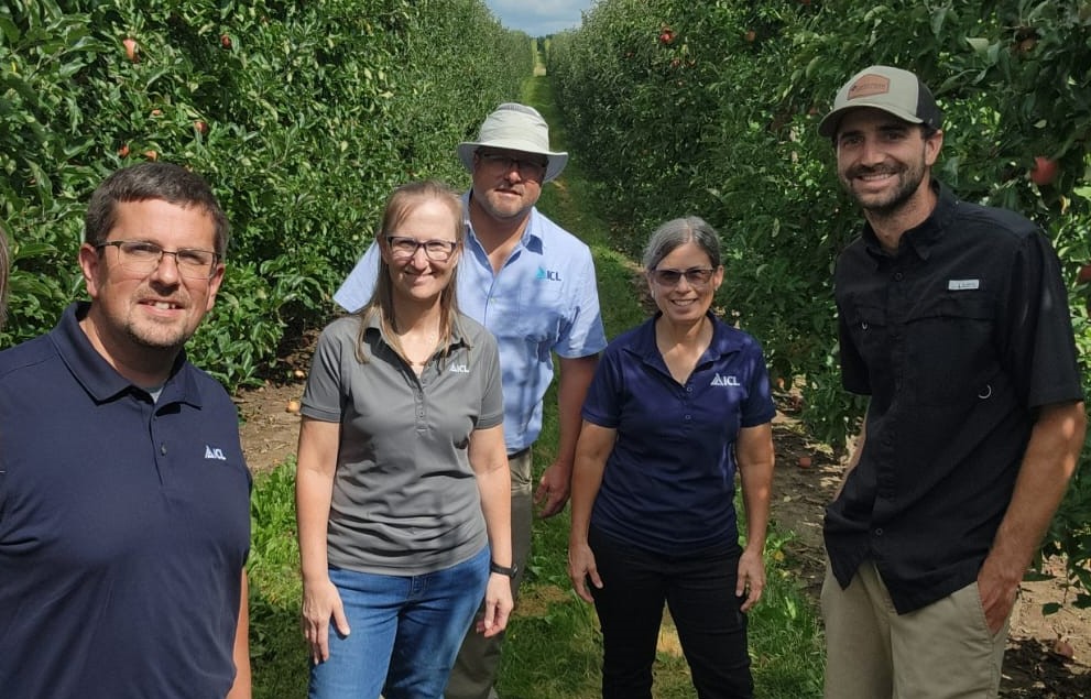 Dr. Jason Haegele and ICL agronomy team standing in an apple orchard during a field visit, surrounded by lush green trees and ripening fruit under clear skies.